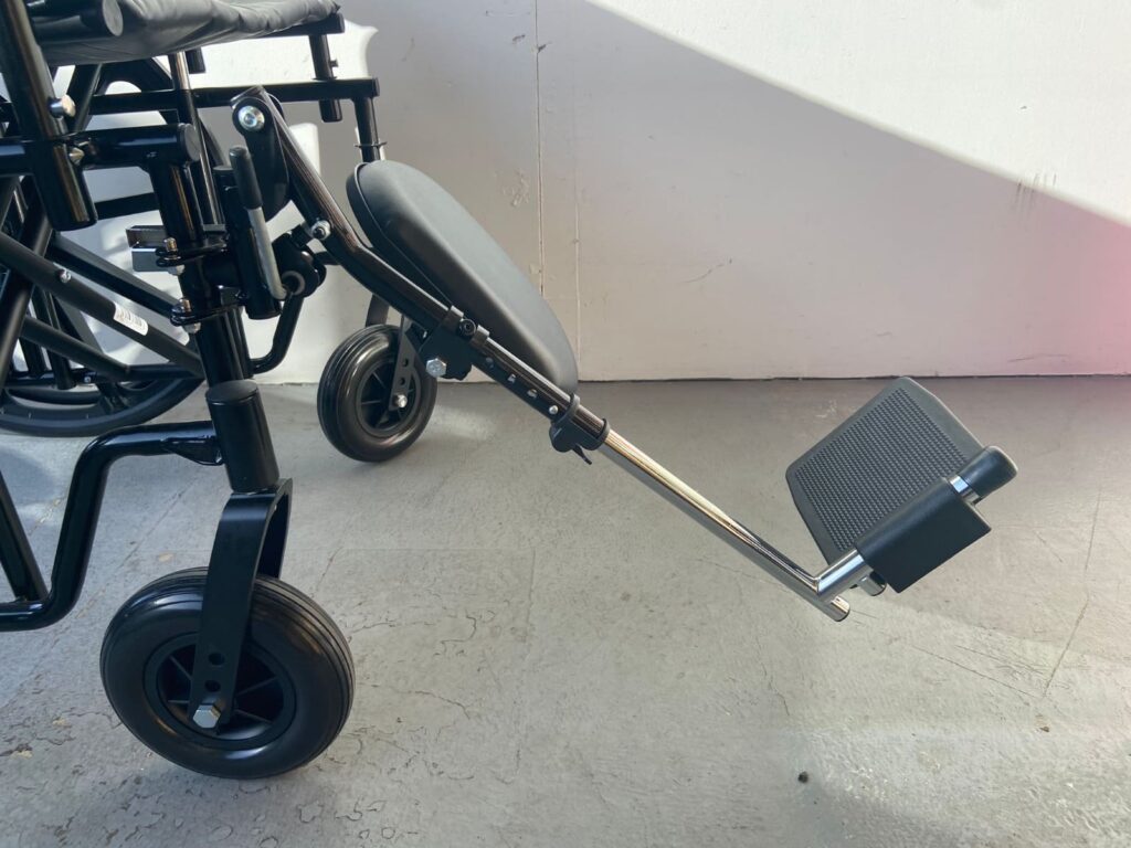 Close-up side view of a black manual wheelchair with an elevated leg rest and footplate extended forward, designed for added comfort and leg support. The image is taken indoors against a light-colored wall with shadows visible.